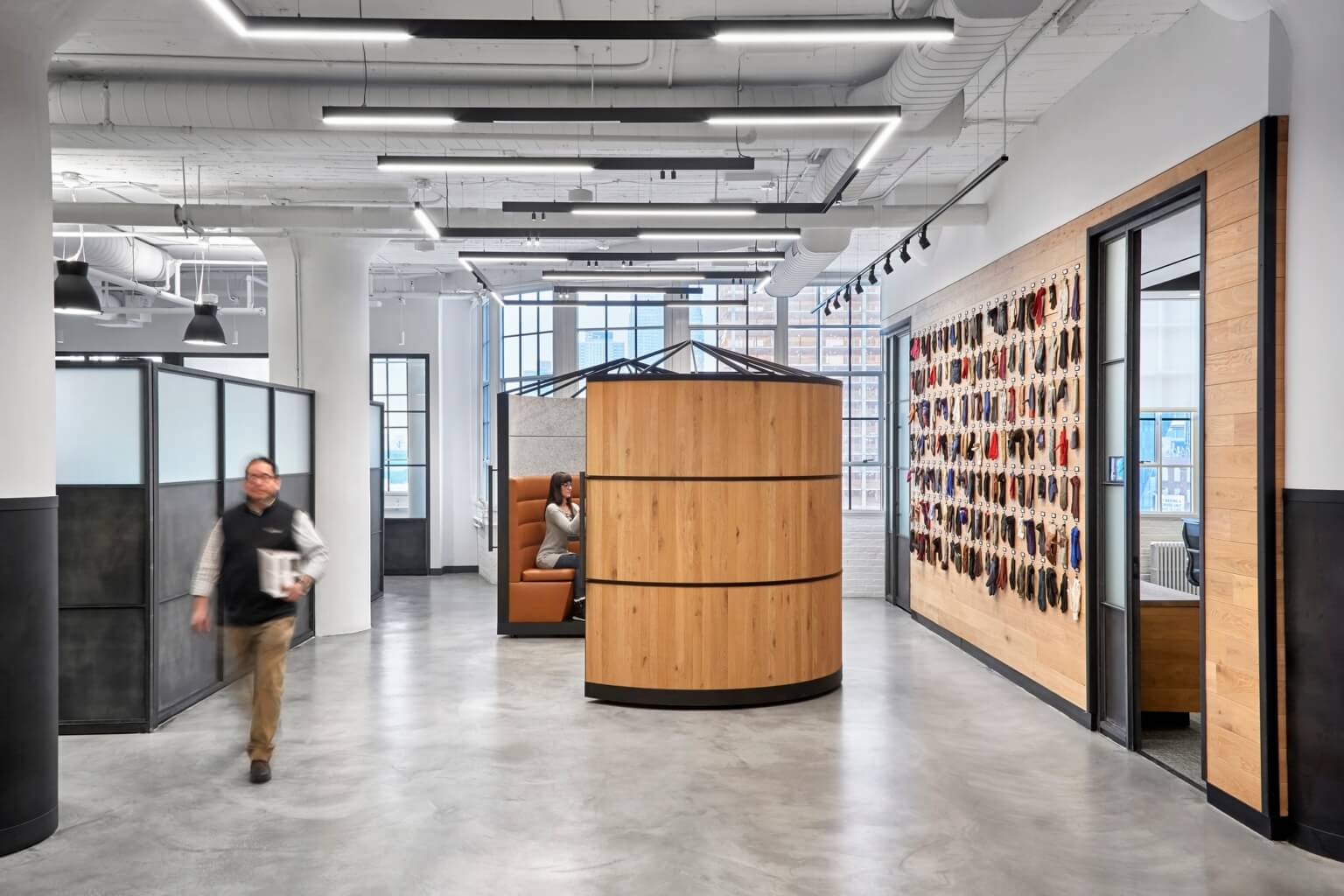 Modern open office space with private seating pods, a wall display of hanging items, and a person walking through the hallway.