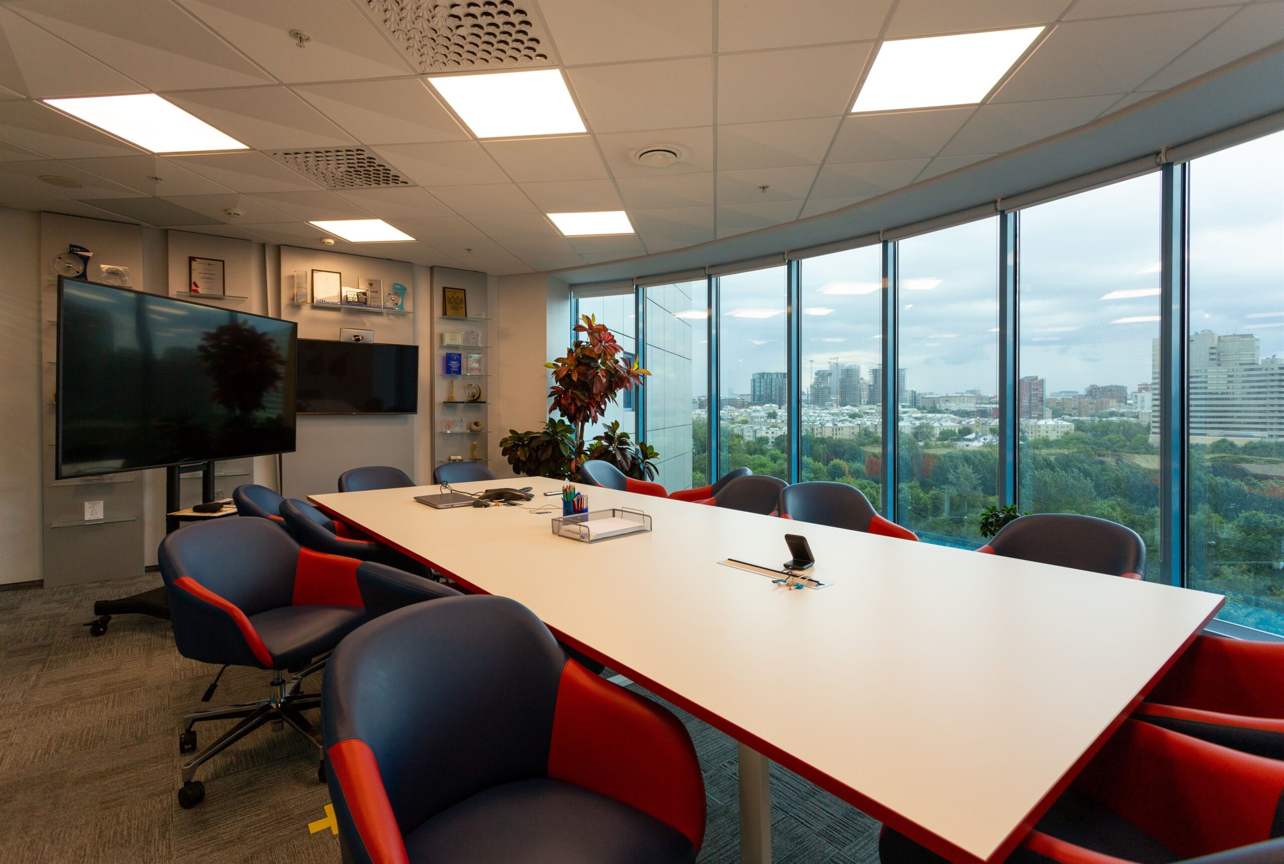 A modern conference room with a long white table surrounded by red and blue chairs.