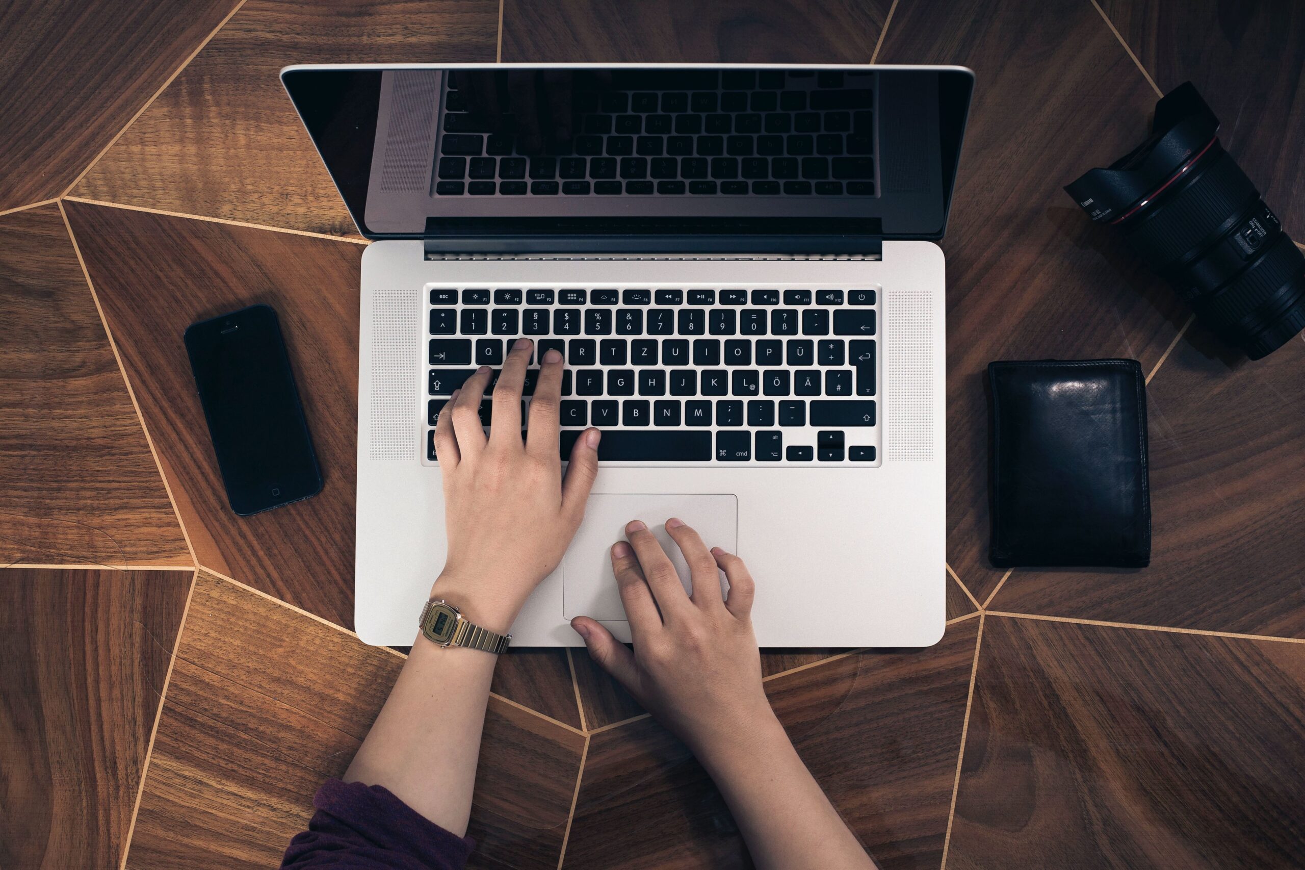 Top-down view of a person using a laptop on a wooden desk, with a phone, wallet, and camera nearby.