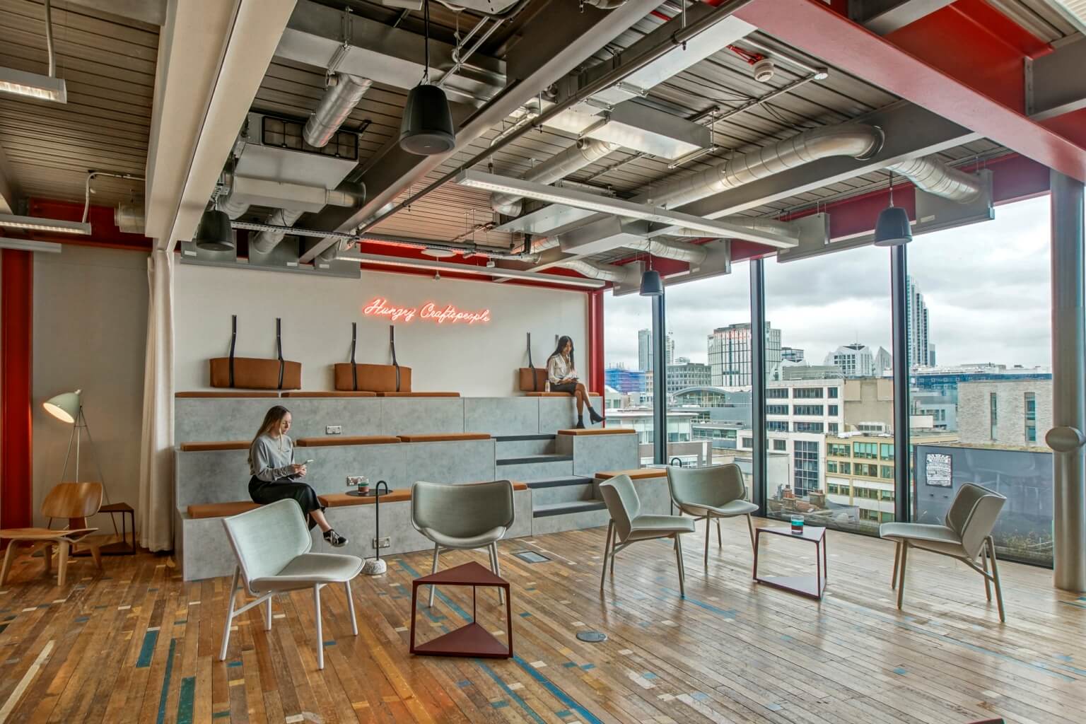 A modern coworking lounge with tiered seating, large windows with a city view, and a neon sign on the wall.