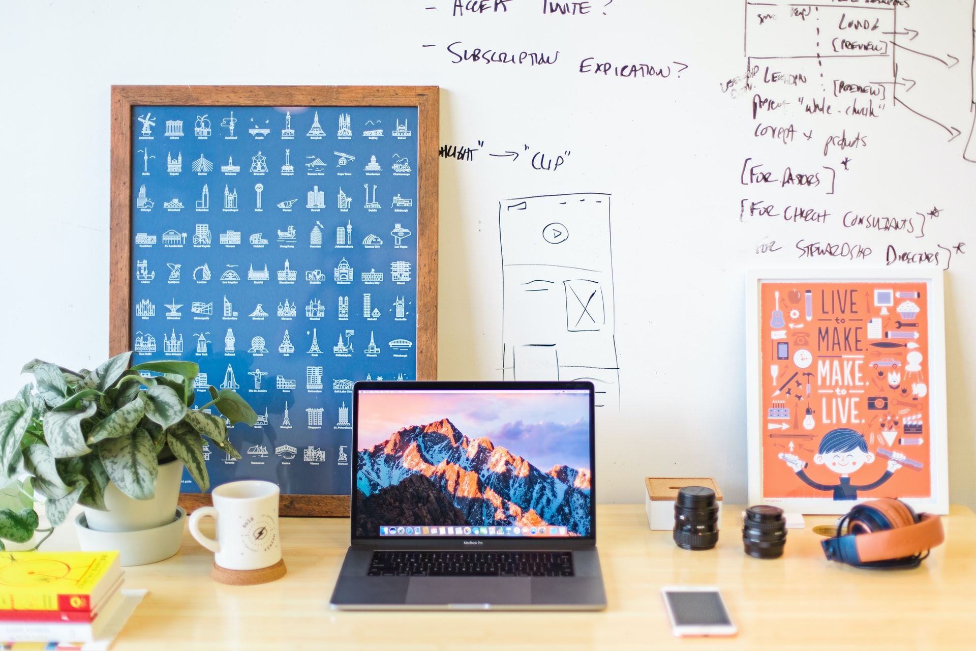 A creative workspace with a laptop on a wooden desk, surrounded by a coffee mug, books, camera lenses, headphones, and framed artwork.