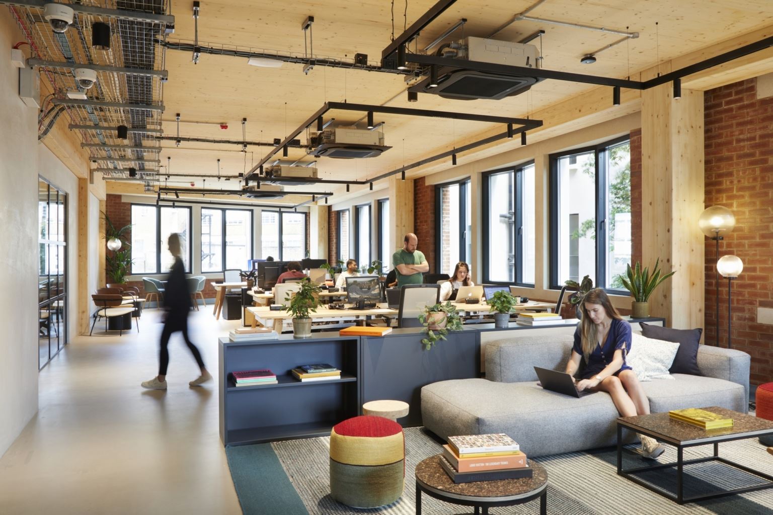 Modern coworking space with people working at desks and on a sofa, surrounded by natural light, plants, and cozy decor.