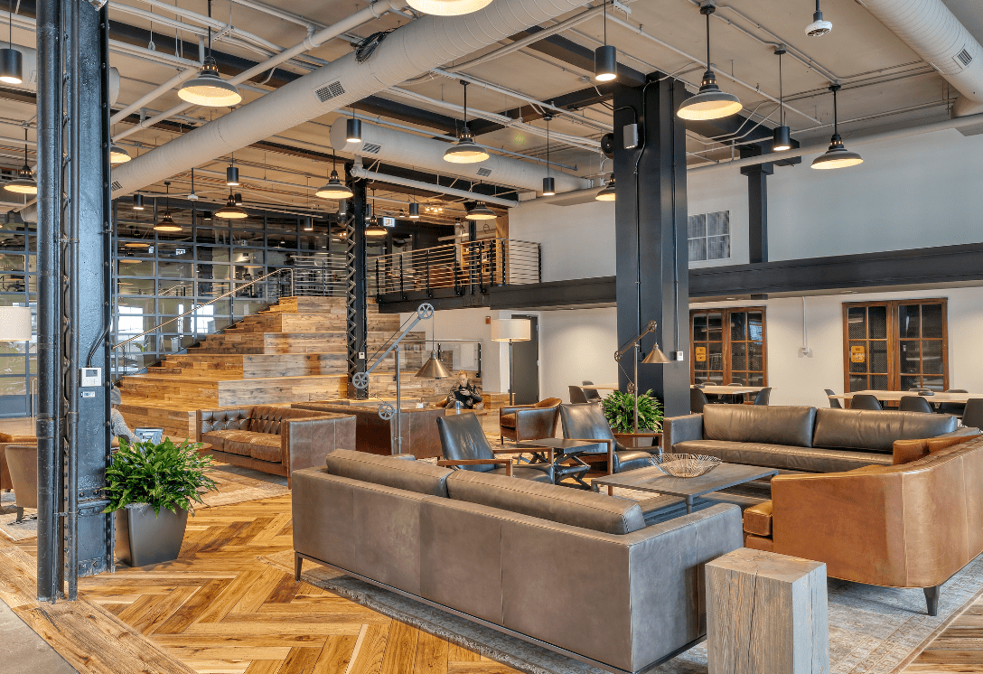 Modern coworking space with leather sofas, wooden floors, open seating areas, and a large staircase leading to an upper level.
