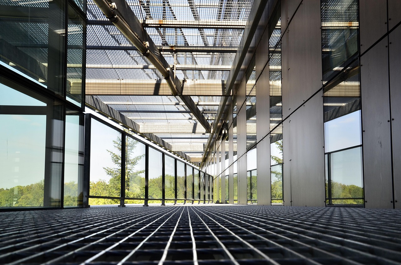 A modern glass walkway with a metal grid floor and overhead metal beams, surrounded by large windows and greenery outside.