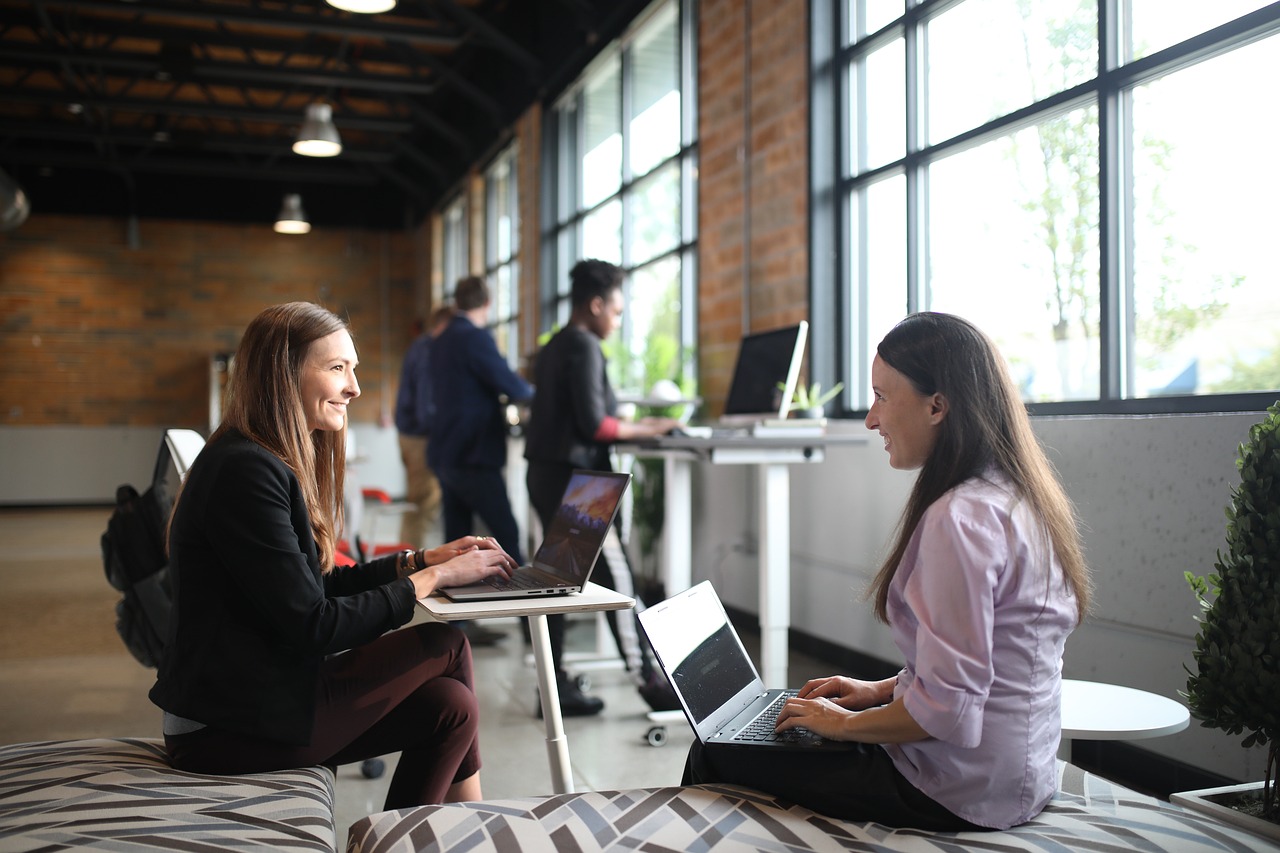 Two women working on laptops and smiling in a modern office space with large windows and a relaxed atmosphere.