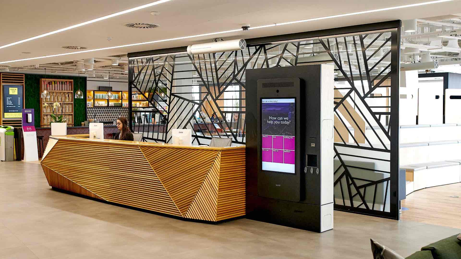 A modern office reception area with a stylish wooden front desk, a receptionist, and a visitor management kiosk made for self-registration.