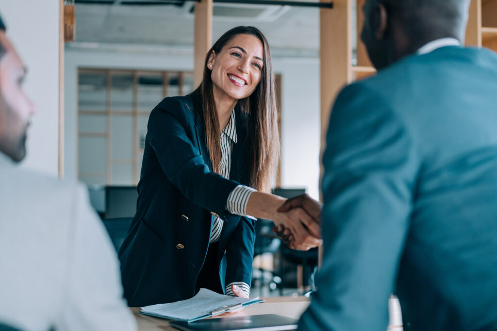 A smiling woman in business attire shakes hands with a man across a desk in a modern office setting.