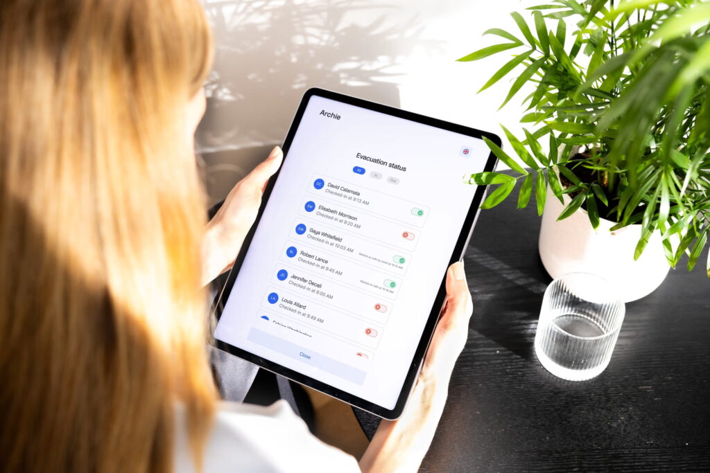 Person holding a tablet showing Archie’s emergency evacuation status screen, next to a glass of water and a potted plant.