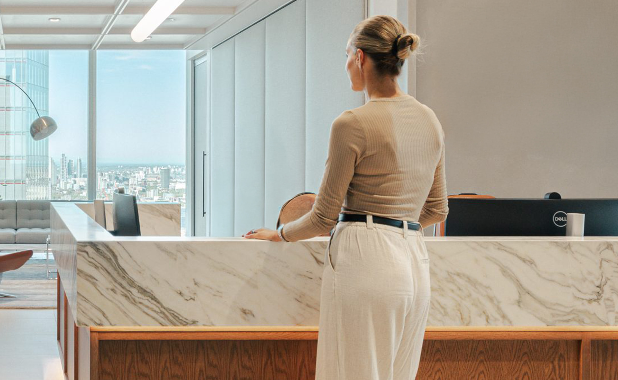 Person standing at a modern office reception desk with a city view in the background.