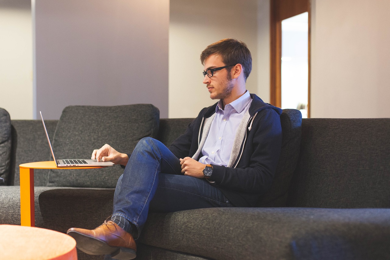A young man in glasses and business casual attire sits on a dark couch, working on his laptop placed on a small orange side table.