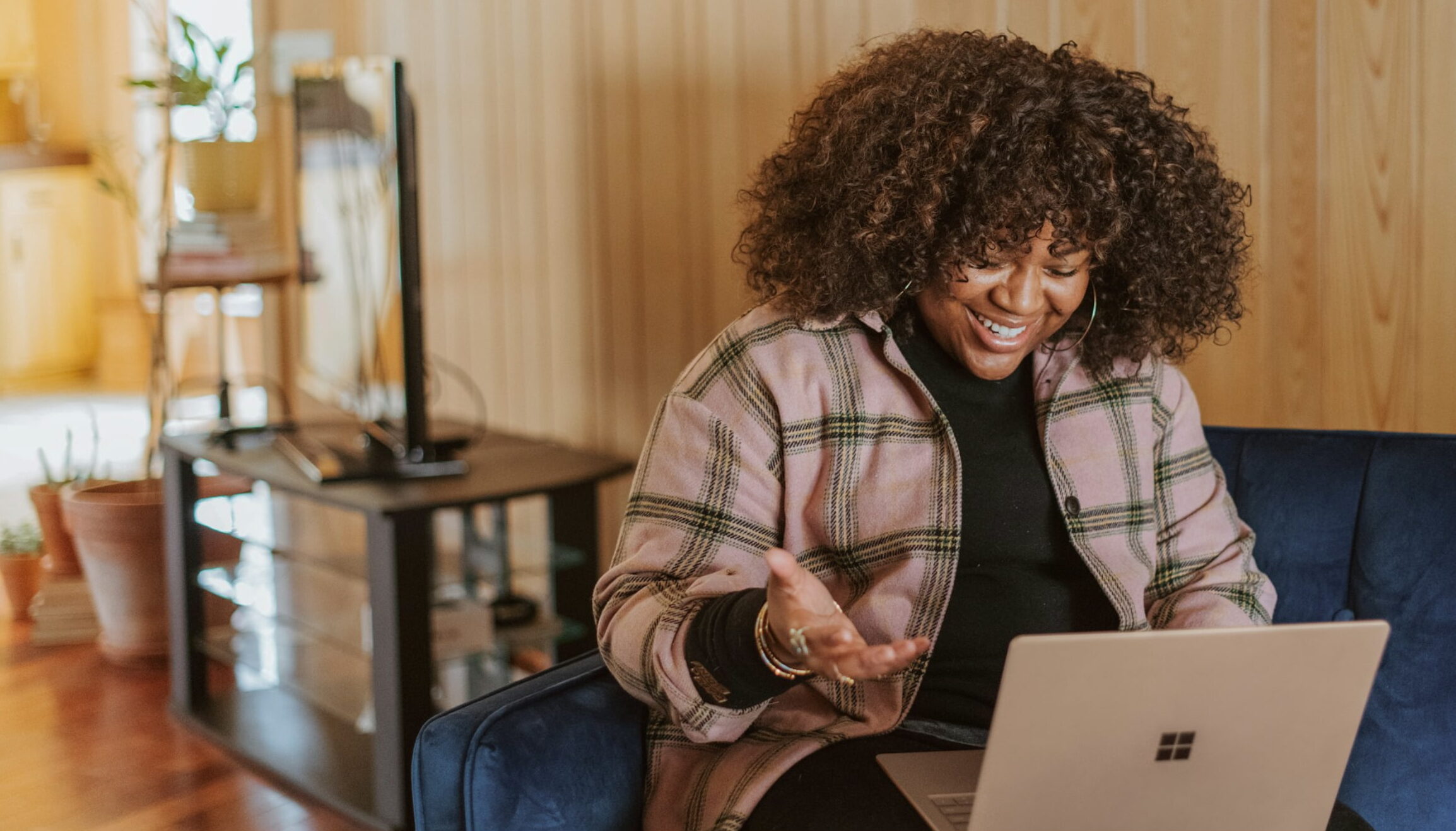 Smiling woman sitting on a couch, video chatting on her laptop in a cozy, wood-paneled room.