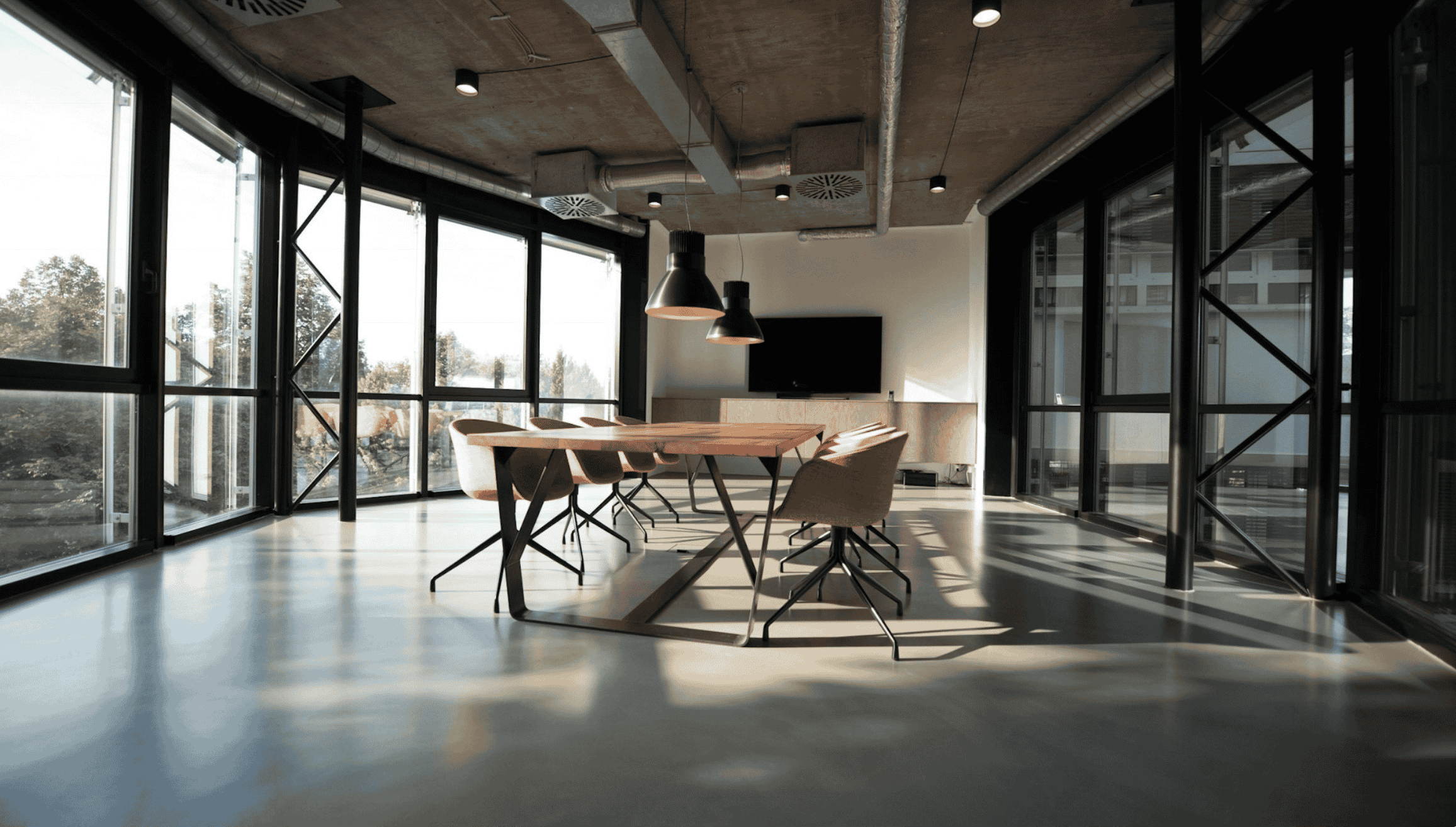 Modern meeting room with large windows, a wooden conference table, pink chairs, and industrial-style ceiling lighting.