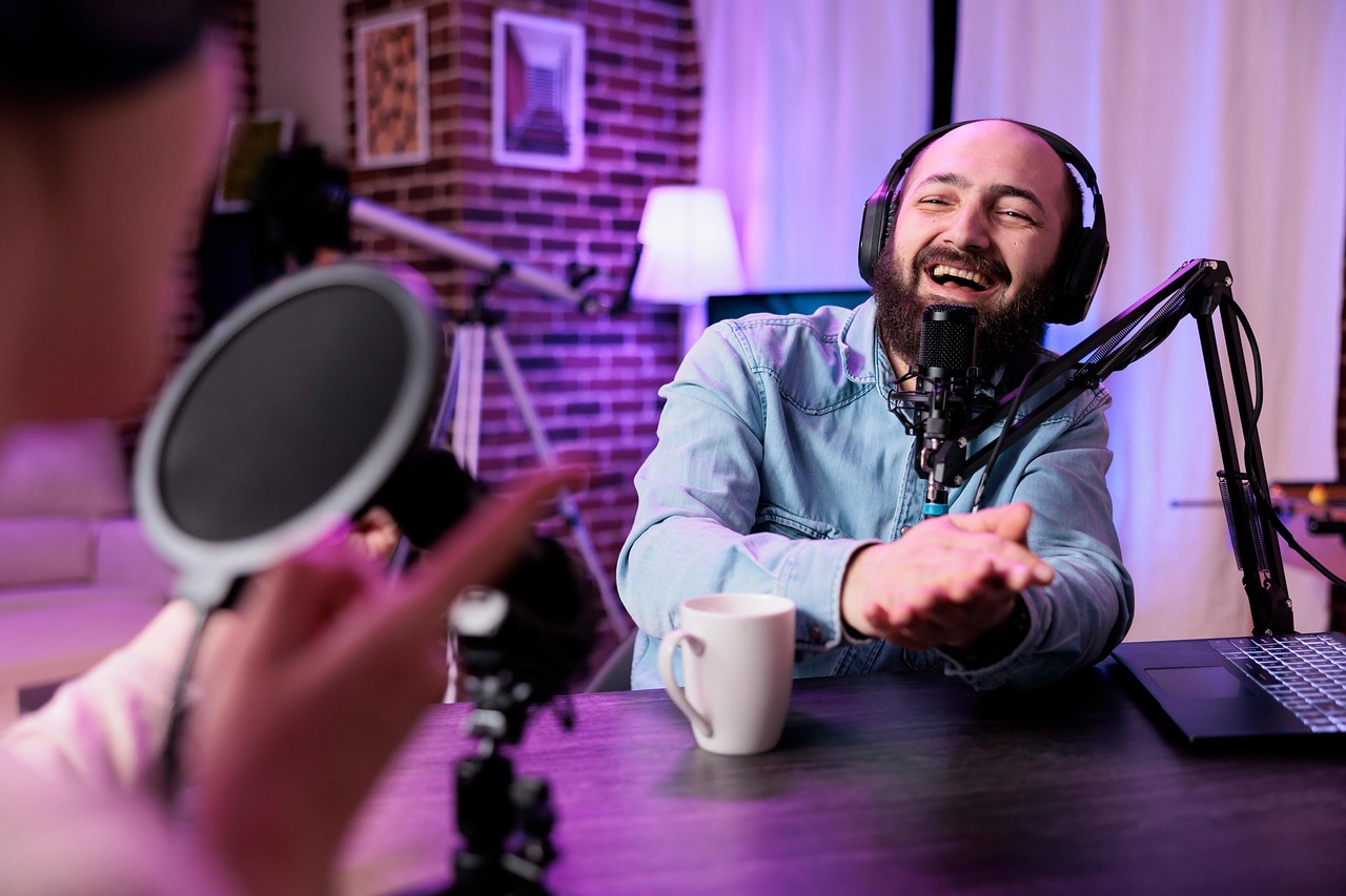 A man recording a podcast in a cozy studio, wearing headphones and speaking into a microphone with a laptop and coffee mug on the table.