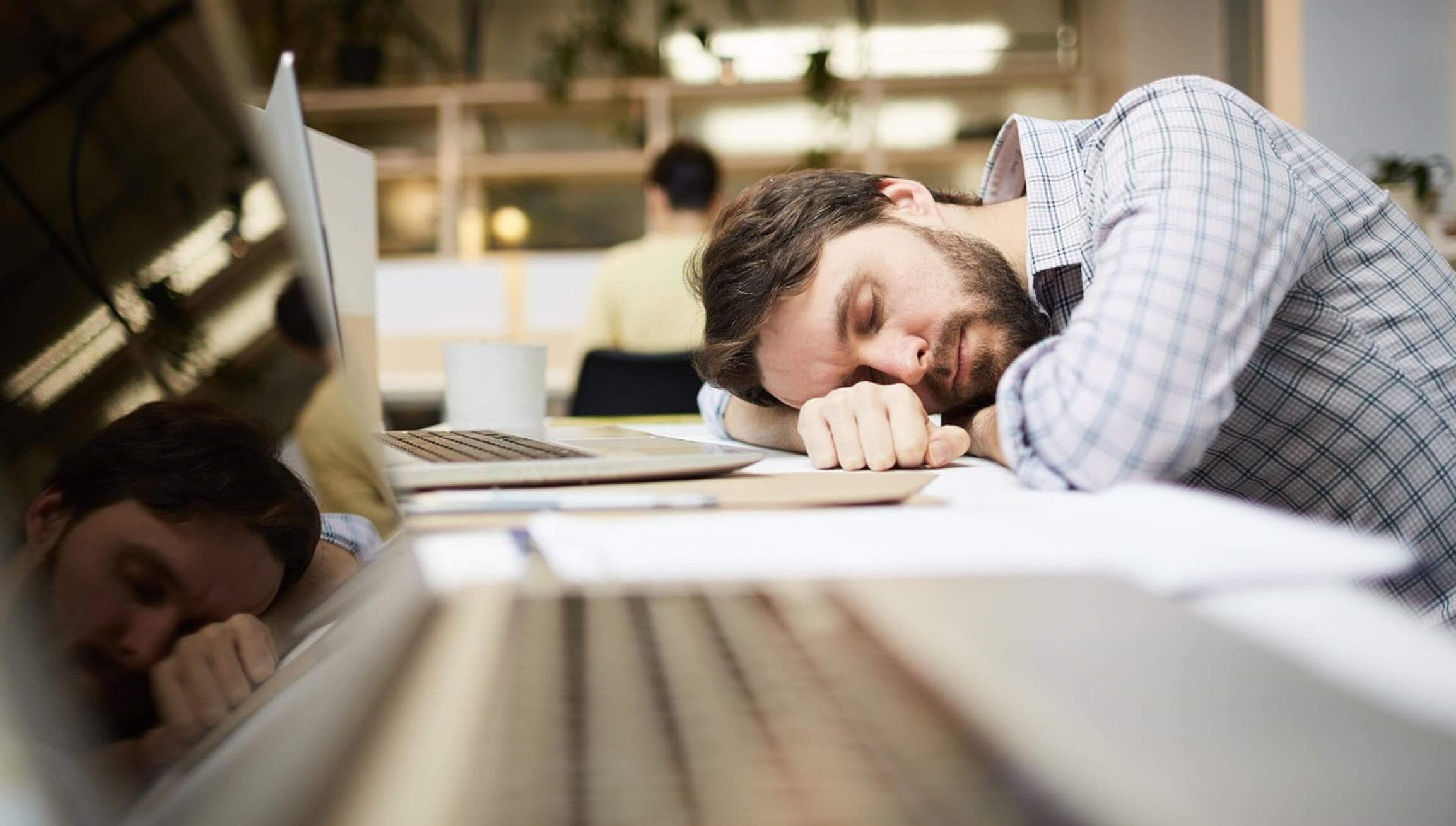 A disengaged employee sleeping at his desk in an office, with his head resting on his arm next to a laptop.