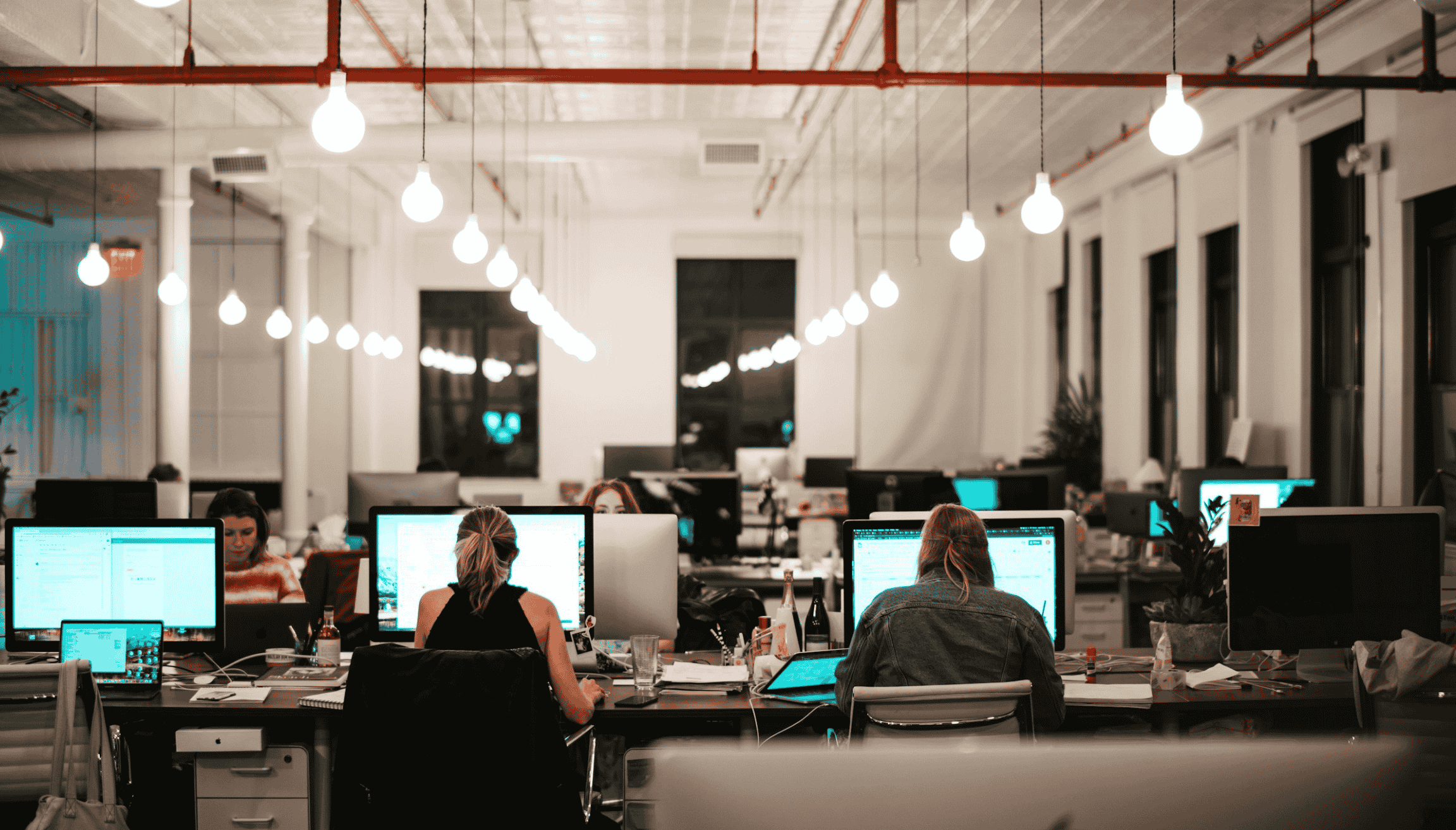 Open-plan office with people working at shared desks, surrounded by monitors and warm hanging lights in a modern coworking space.