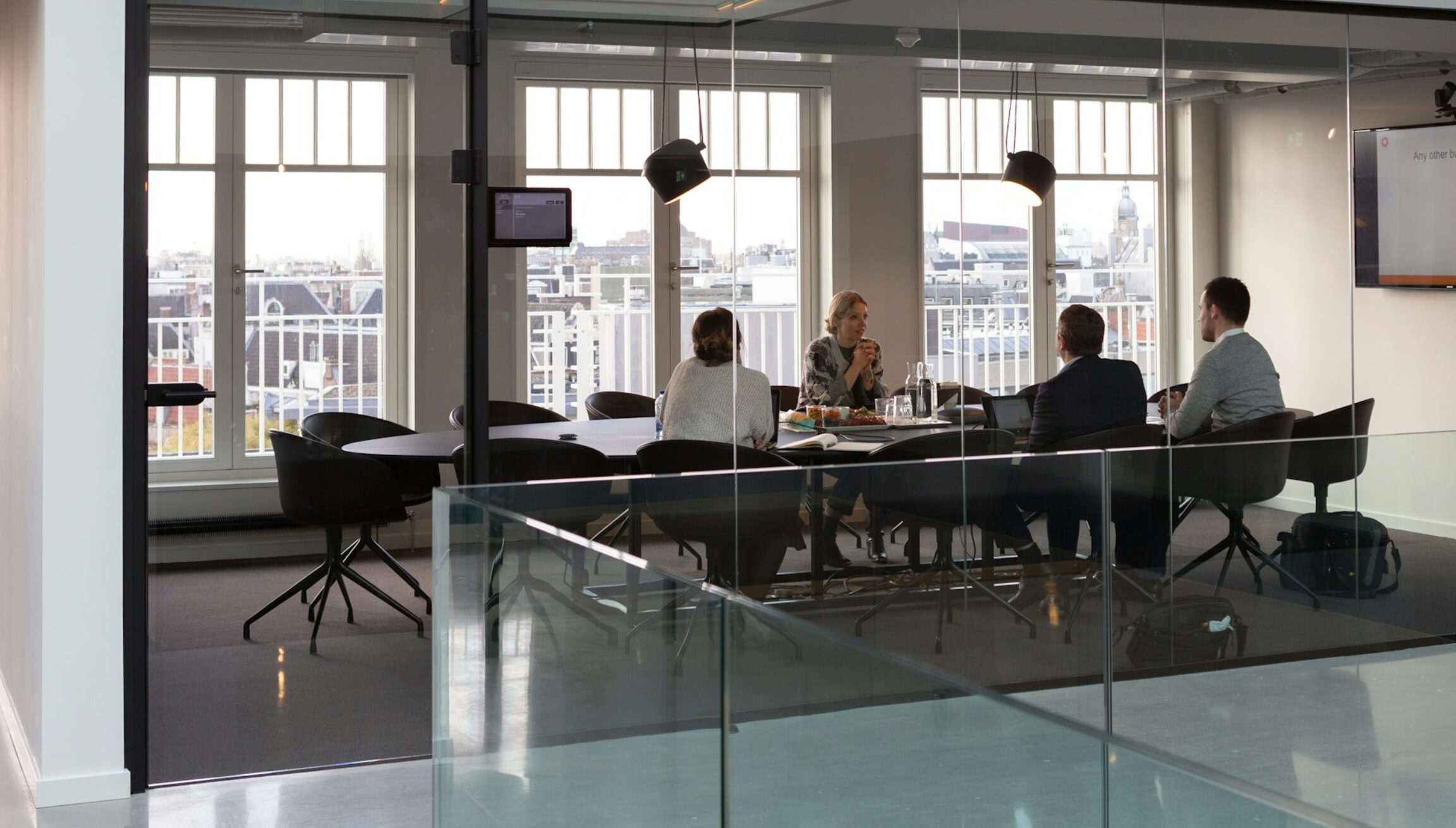Four professionals in a glass-walled meeting room having a discussion, with city views in the background.