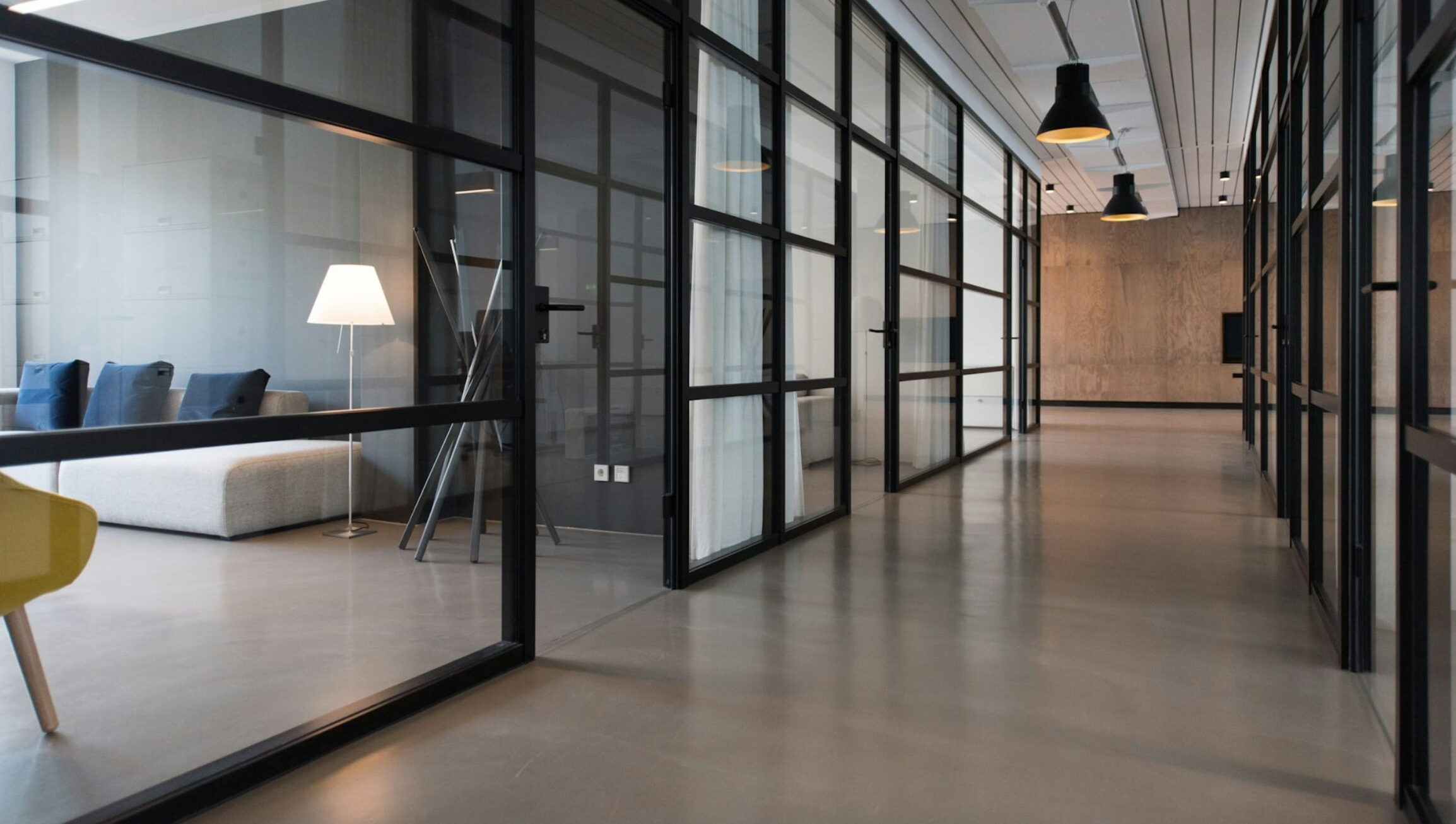 A modern office hallway with glass-walled meeting rooms, minimalist furniture, and overhead lighting.