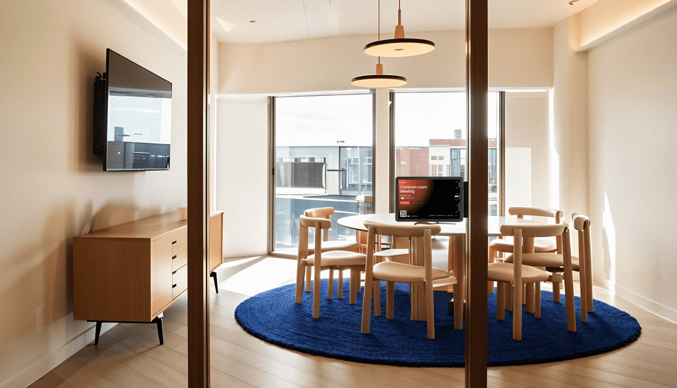 Small modern meeting room with wooden chairs around a round table, digital room display showing booking details, and large windows with city views.