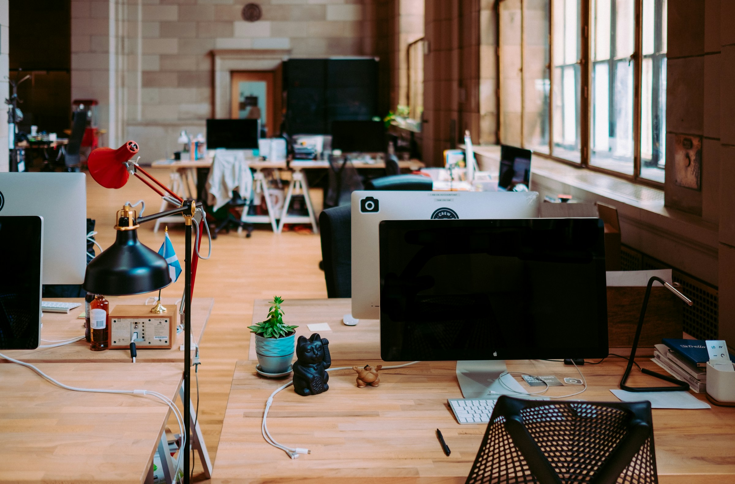 Open-plan office with desks, iMacs, desk lamps, and natural light from large windows.