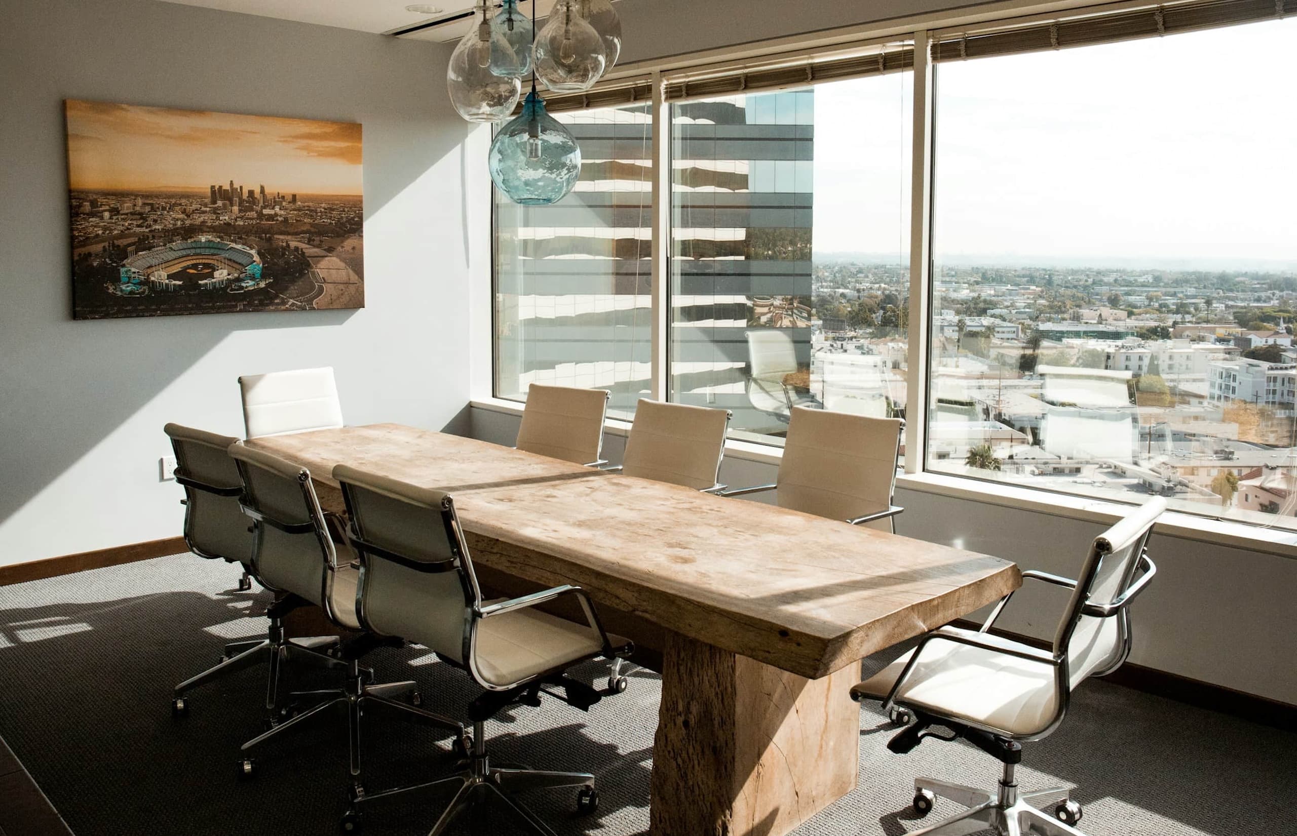 Empty meeting room with a large wooden table, white office chairs, and floor-to-ceiling windows overlooking a cityscape.
