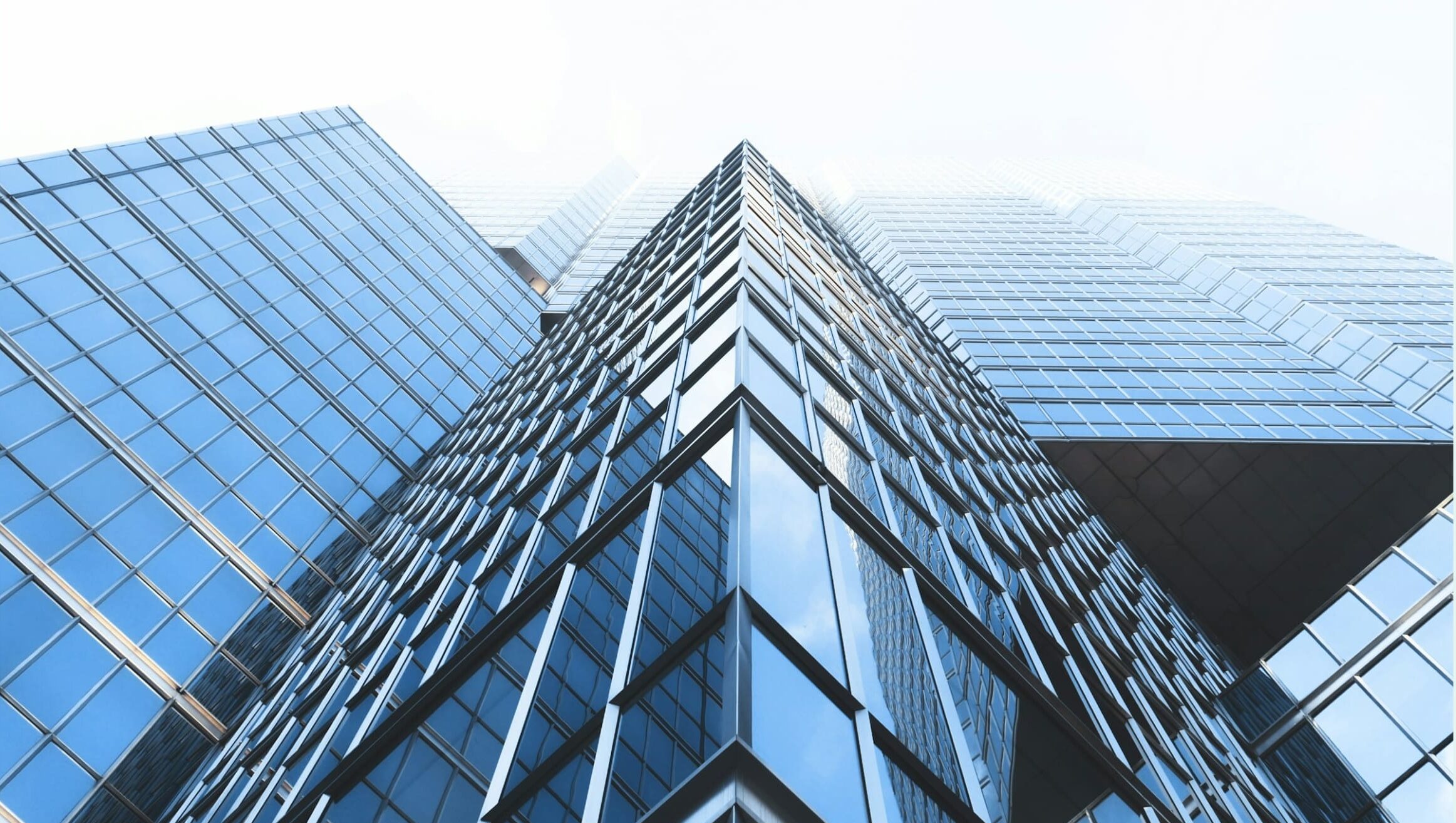Low-angle view of a modern glass skyscraper with reflective windows and sharp architectural lines against a bright sky.