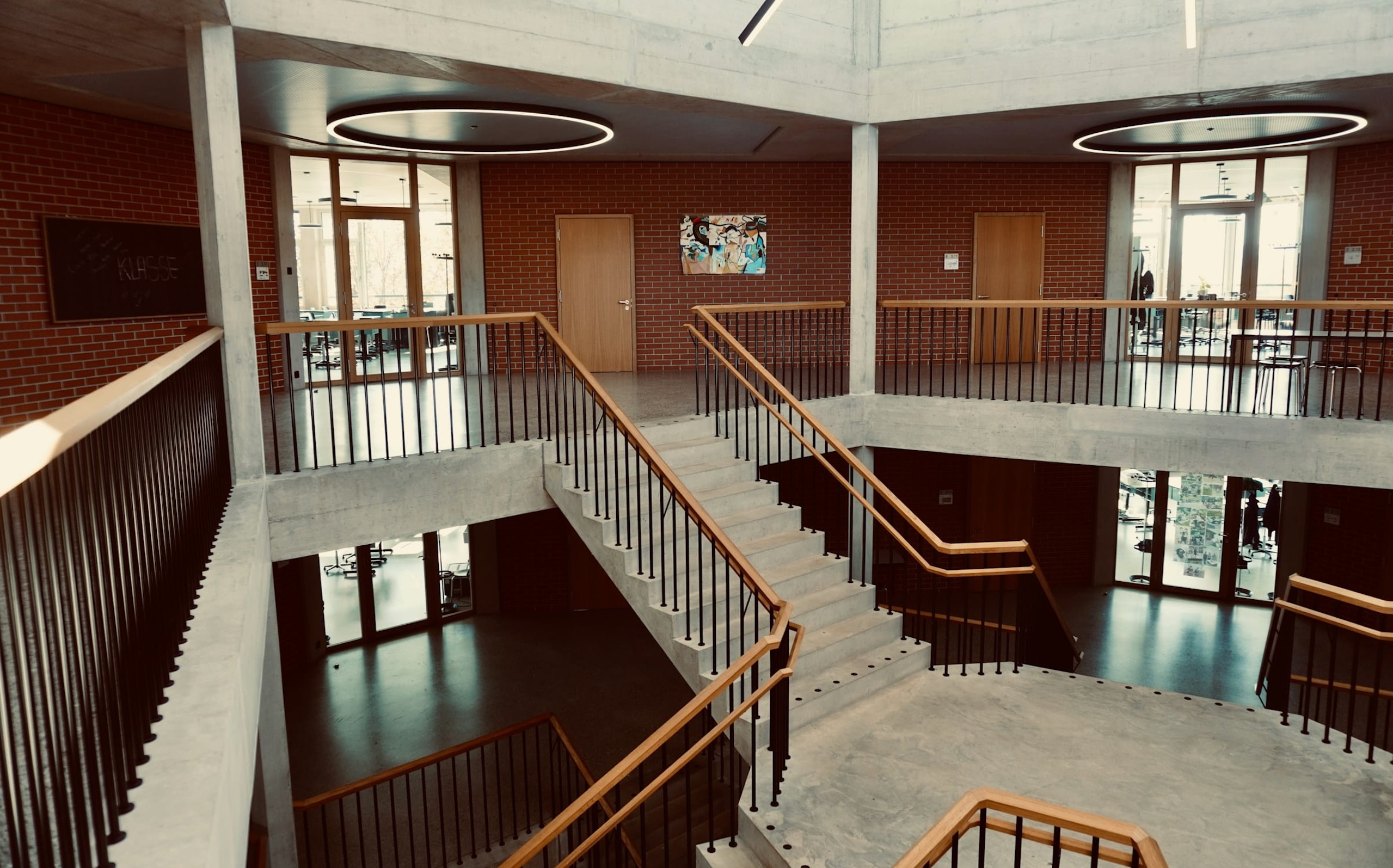 Interior of a modern building with exposed concrete, brick walls, and a central staircase surrounded by glass doors and classrooms.