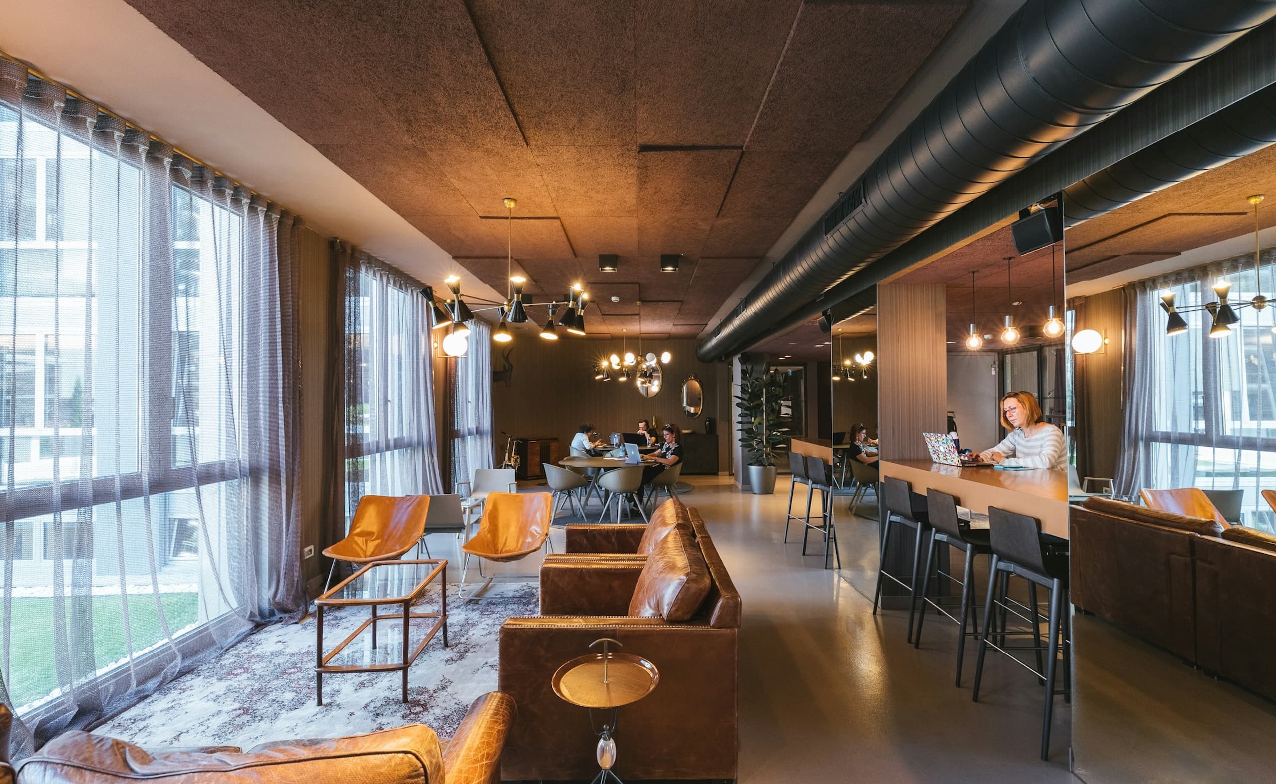 Modern coworking lounge with large windows, cozy seating, and people working at shared tables and bar-height desks.