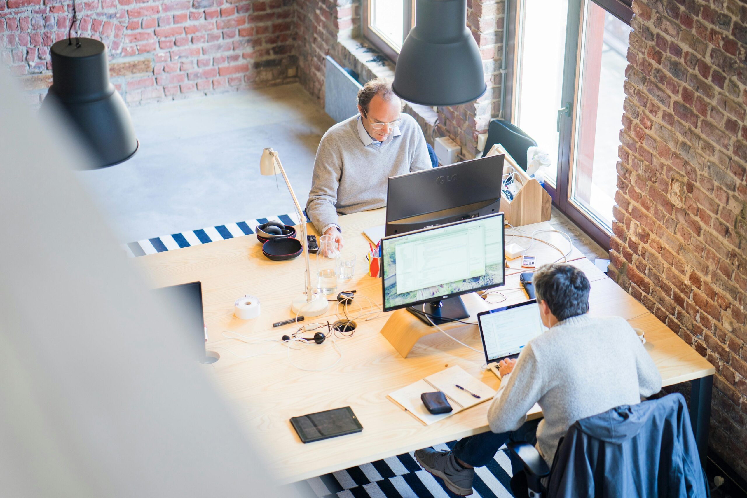 Two people working at a shared wooden desk in a bright, brick-walled office with striped rugs and large black pendant lamps.