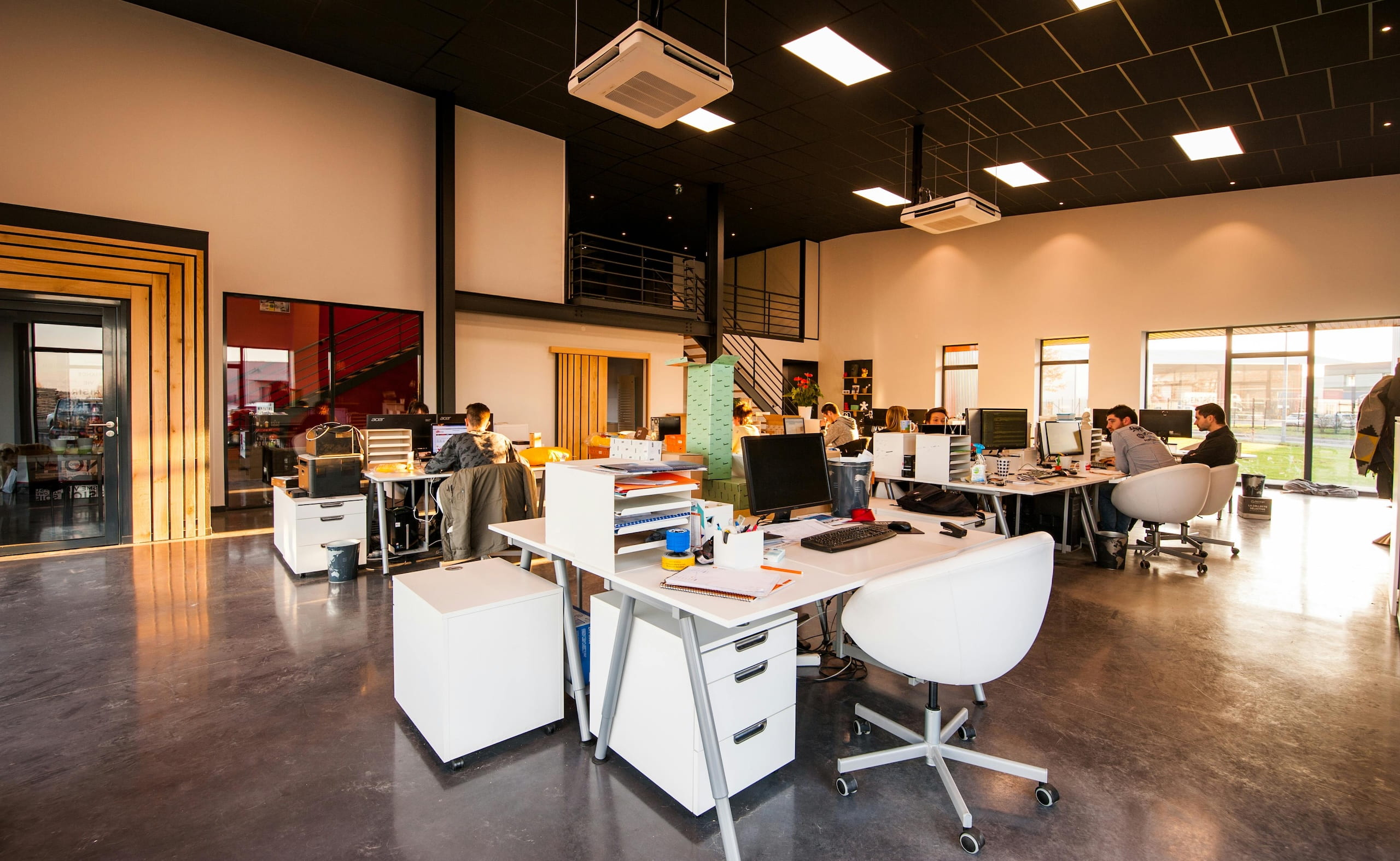 Open-plan modern office with white desks and chairs, where teams are working on computers in a bright space with large windows.