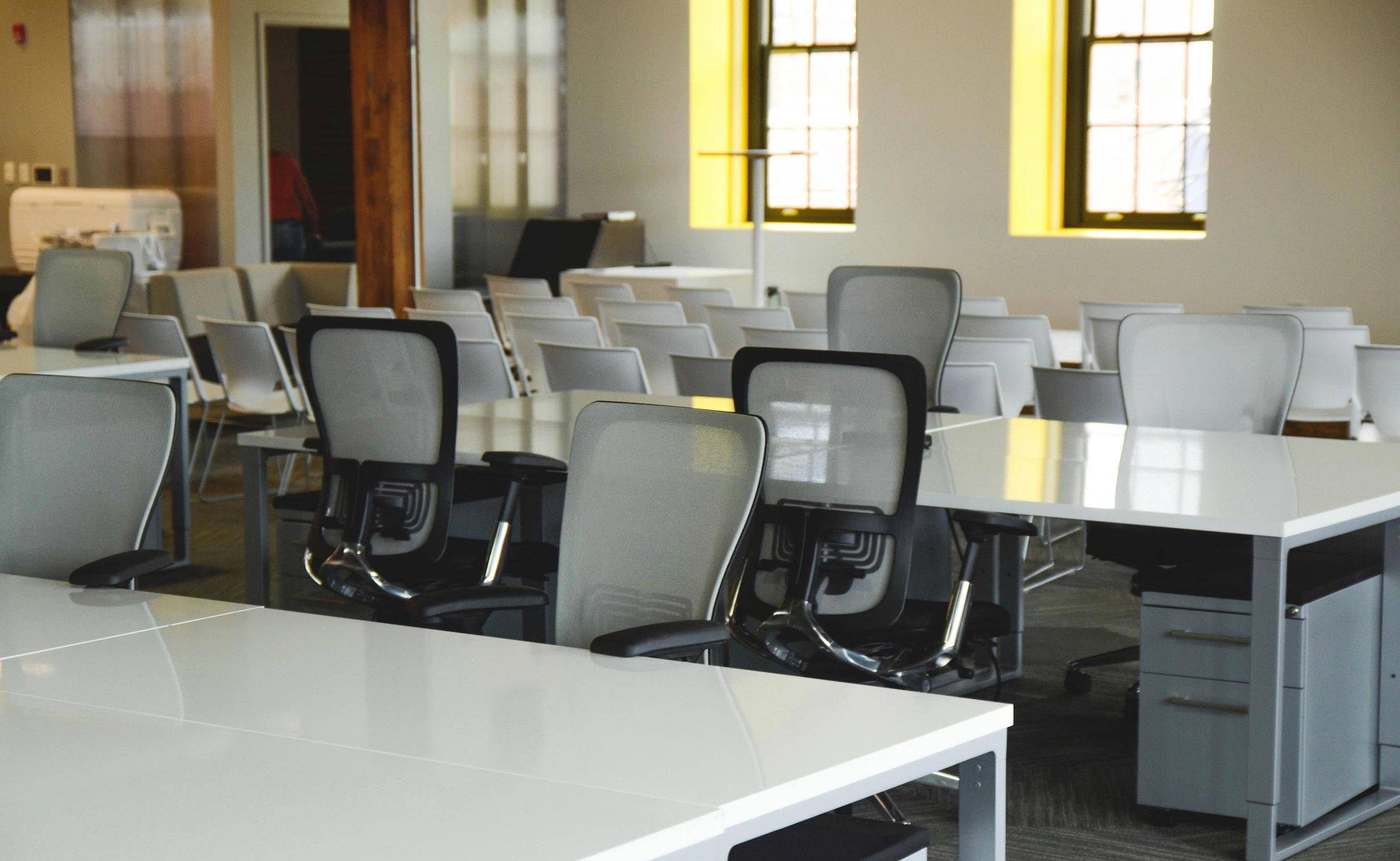 Bright coworking space with white desks, ergonomic chairs, and natural light coming through large windows.