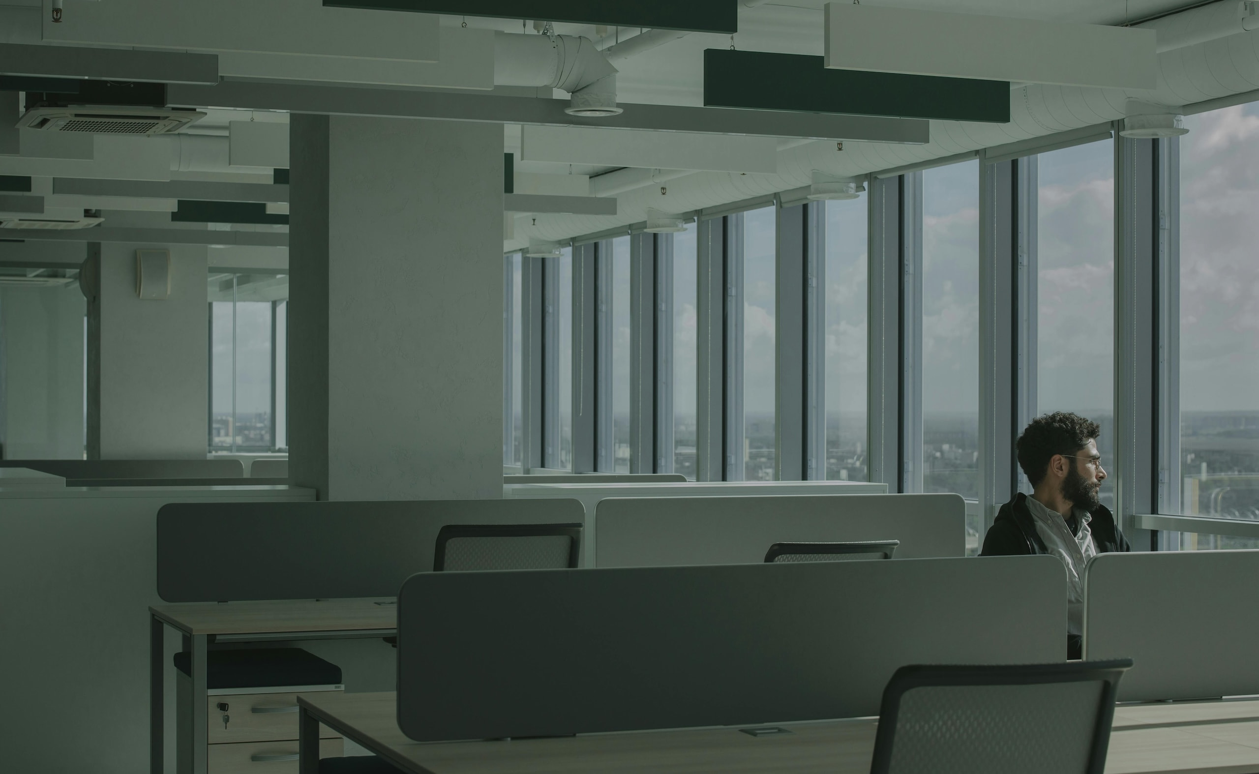 Quiet open office with desk dividers and a person sitting by large windows overlooking the city.